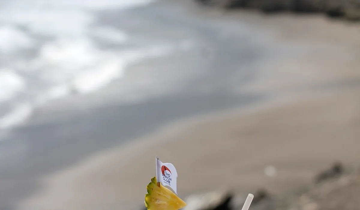 Close-up of a hand holding a yellow object with a small tag, with a blurred beach and ocean in the background.