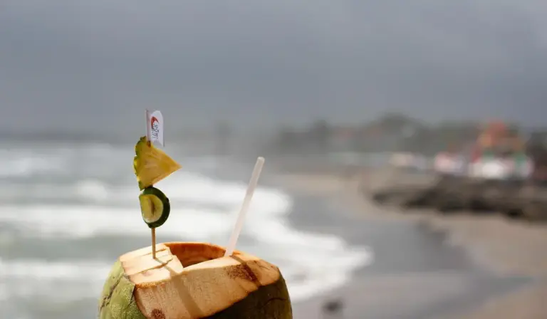 A coconut drink with a straw and lime wedge on a sandy beach with the ocean and cloudy sky in the background.
