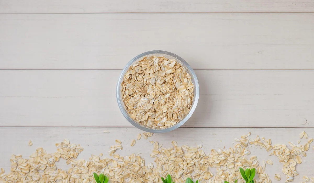 Bowl of rolled oats in a small dish on a pale wooden surface with scattered oats nearby.