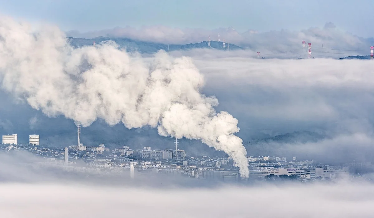 Industrial smokestacks releasing white smoke over a city shrouded in clouds.