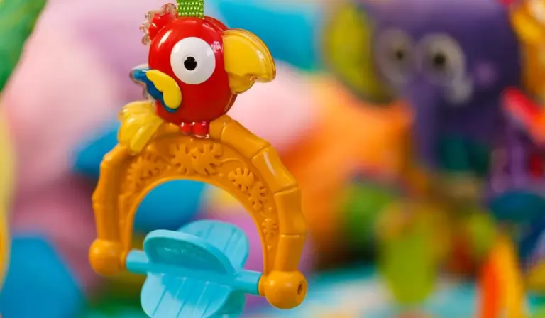 Bright red parrot-shaped chew toy perched on a yellow ring with blue blades, set against a colorful, blurred background.