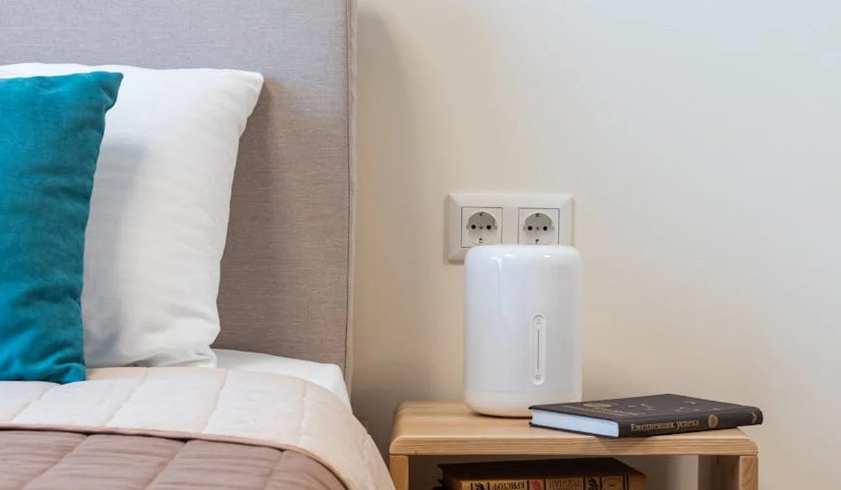 A compact humidifier sits on a wooden nightstand beside a bed with pillows and a book, illustrating a calm setup for humidity control in a parrot's living space.
