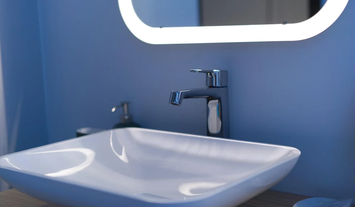 White ceramic bathroom sink with a chrome faucet mounted on a blue wall, with a round backlit mirror above.