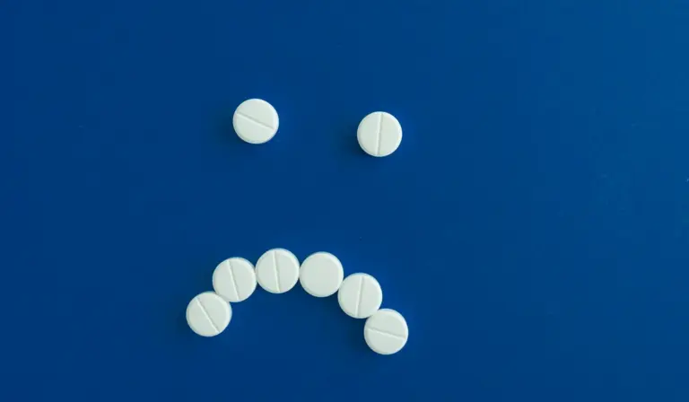 Blue background with white pills arranged to form a sad face (two pills as eyes and a curved row as a mouth).