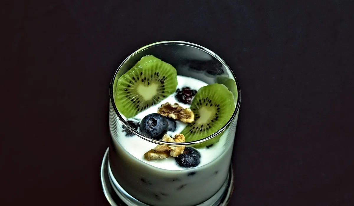 A glass of creamy yogurt topped with sliced kiwi, blueberries, and a sprinkle of granola.