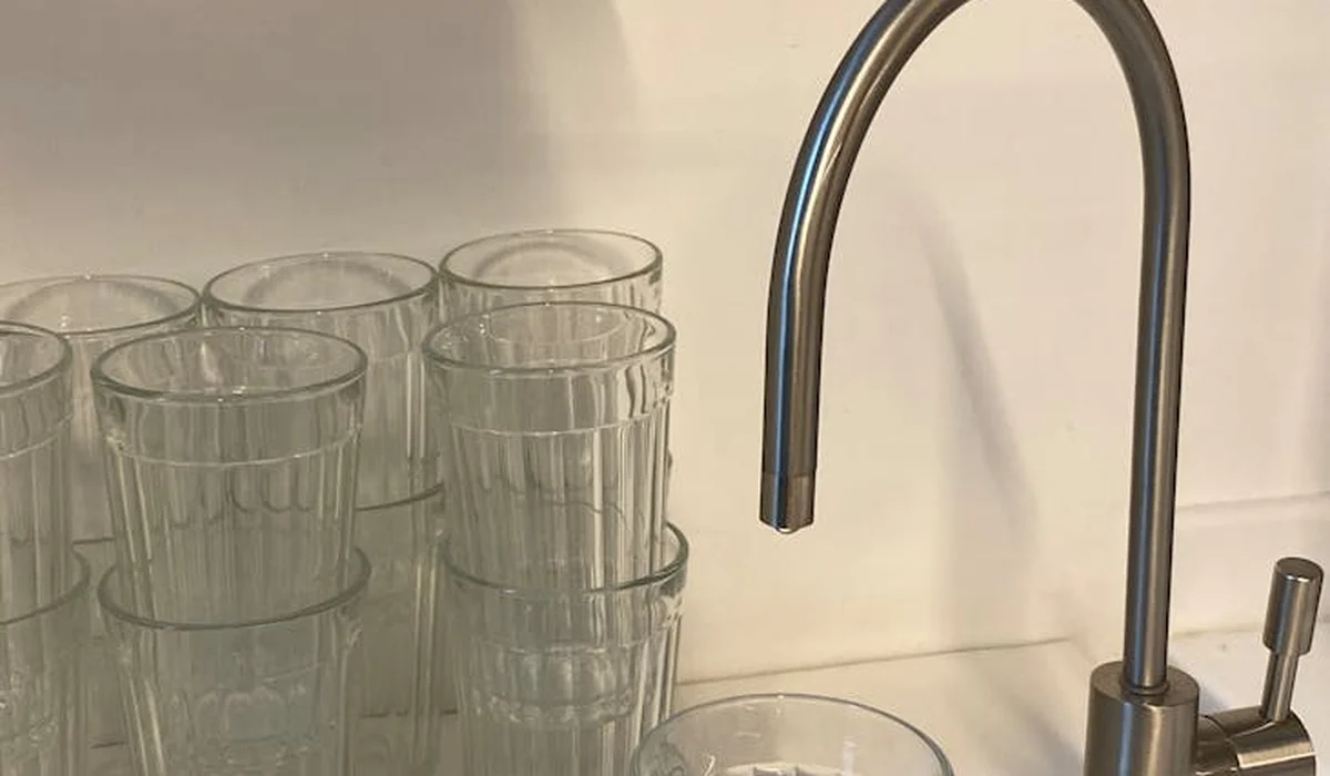 Close-up of a kitchen sink with a tall curved faucet and a stack of clear drinking glasses, suggesting a prepared water-use setup for parrots.