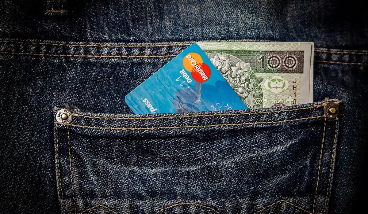 Close-up of a denim jeans pocket with a blue credit card partially visible and cash peeking out.