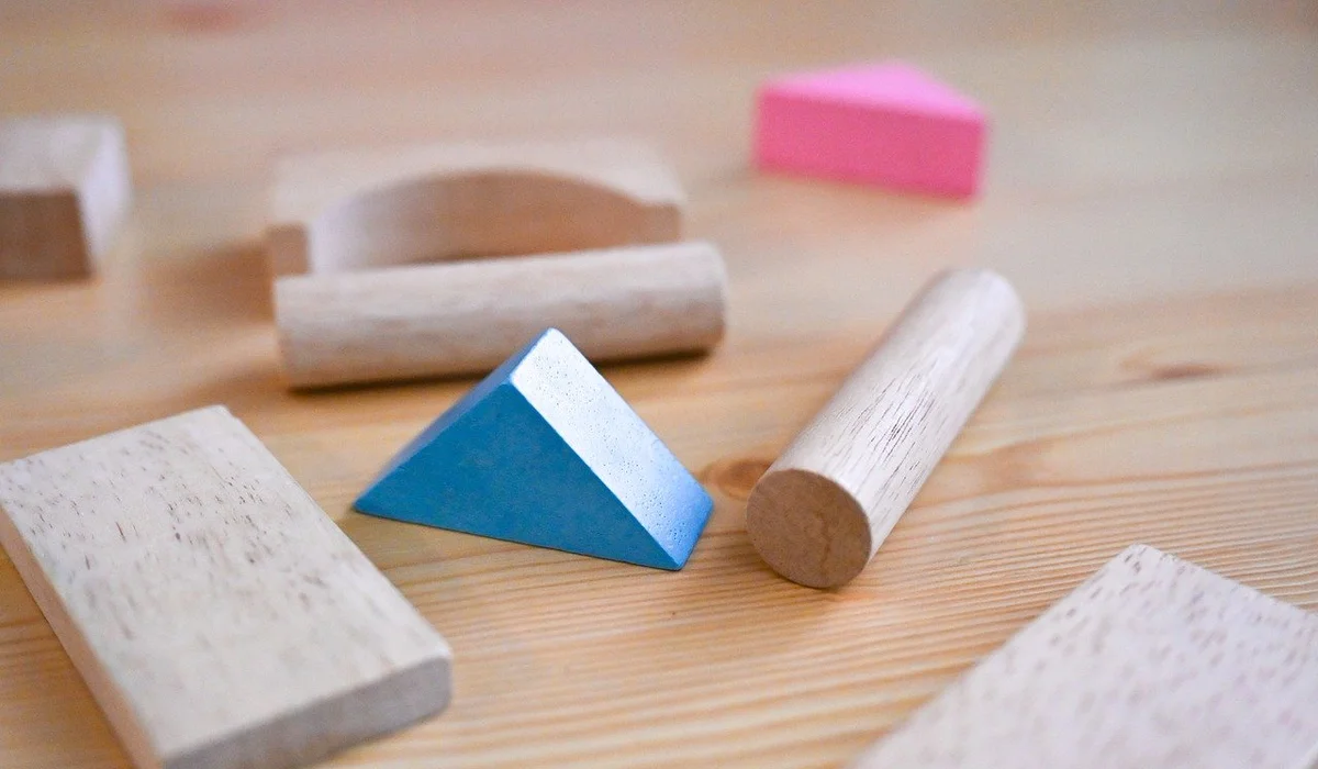 Close-up of assorted wooden toy blocks and shapes on a wooden surface, including a blue triangular prism, a rounded cylinder, a rectangular block, and a pink cube.
